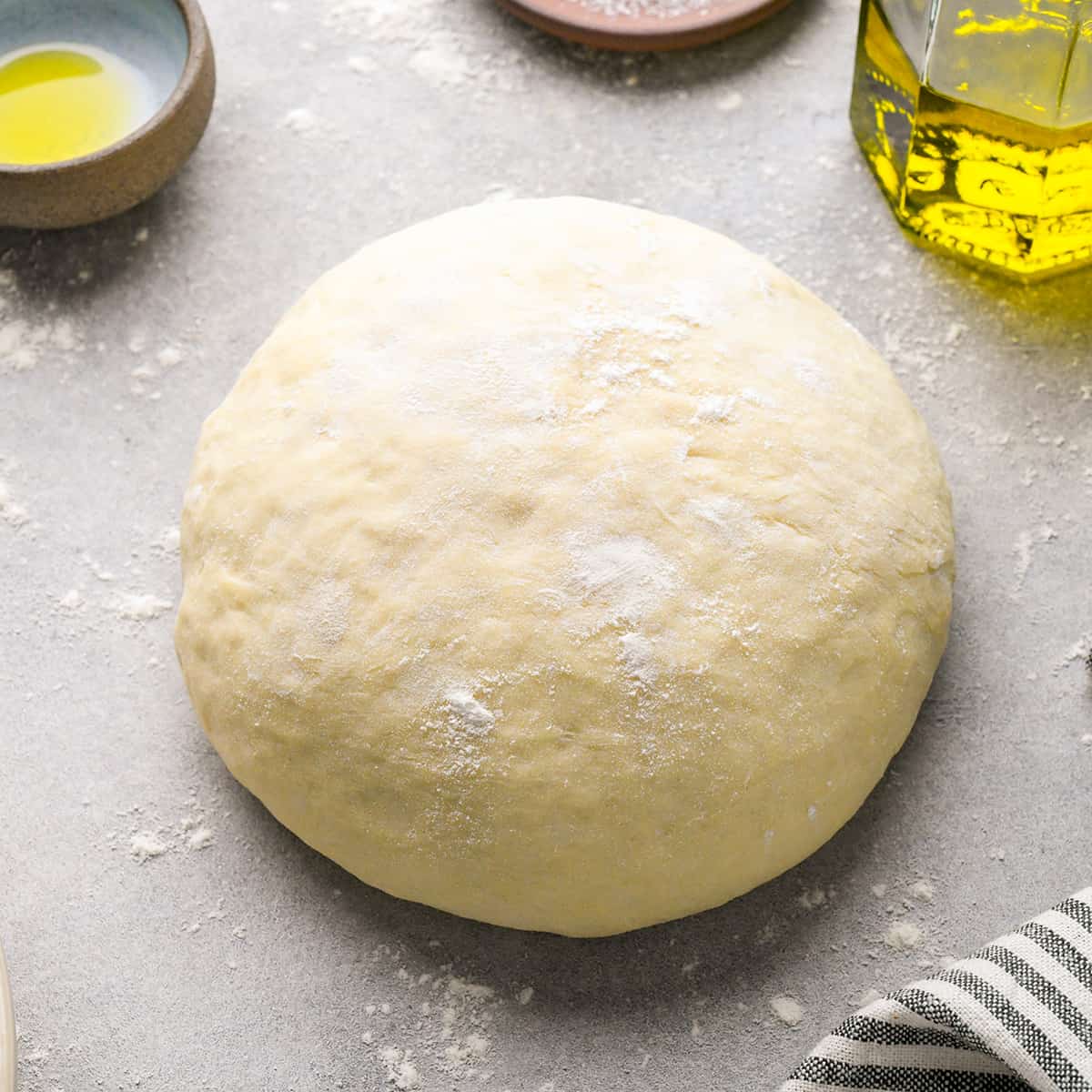 This Easy Homemade Pizza Dough recipe yields the best ever homemade pizza in 30 minutes FLAT! It’s made with only 5 ingredients and does not require any rising! Plus it is vegan with a gluten-free option! #pizzadough #vegan #glutenfree #homemade #easy #norising #homeamdepizza #pizza a ball of homemade pizza dough dusted with flour on a gray floured surface with olive oil and a striped towel nearby