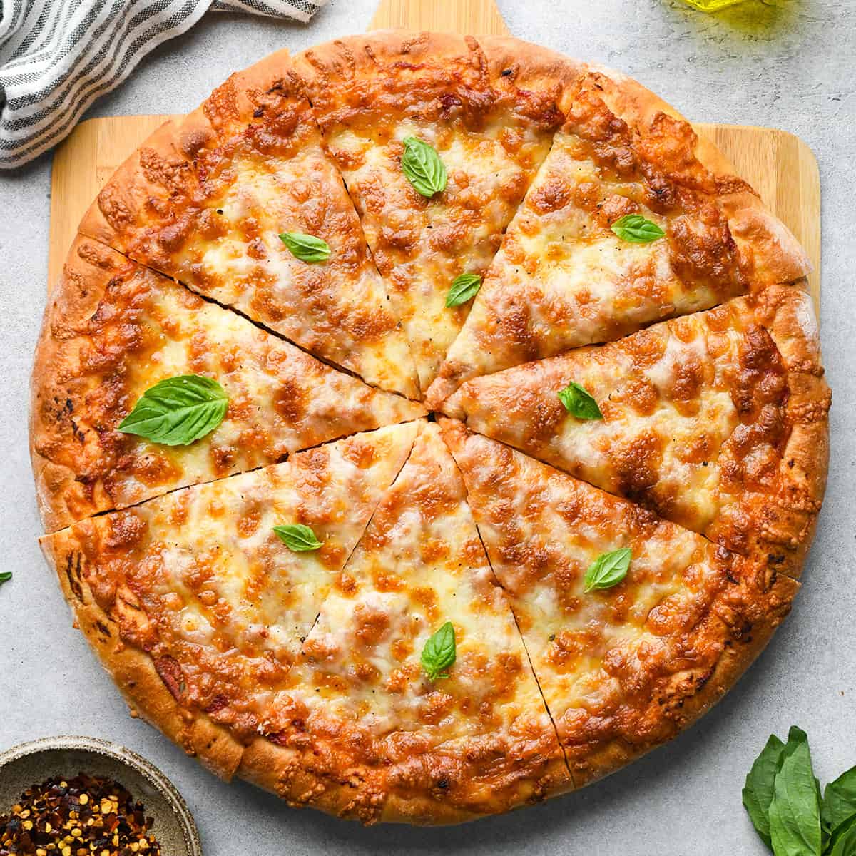 a cheese pizza made with this pizza dough recipe garnished with fresh basil and cut into 8 triangular slices. There is a striped towel, red pepper flakes, fresh basil and olive oil nearby.